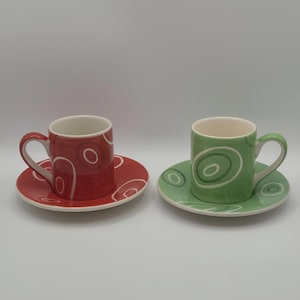 May include: Two ceramic espresso cups and saucers. One is red with white swirl designs, the other is green with white swirls. Both have white interiors and matching saucers. The cups have curved handles.