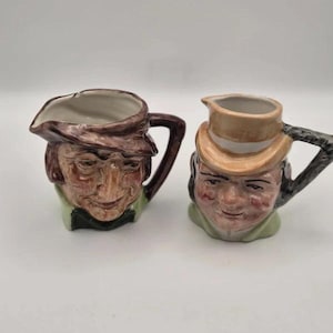 May include: Two ceramic character jugs shaped as human heads with handles. The left jug features a person in a brown hat with a green collar. The right jug depicts a person in a tan top hat with a green collar. Both have painted facial features.