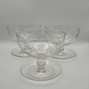 May include: A set of three clear glass coupes, each featuring a shallow bowl with a delicate etched star pattern, a short stem with a central spherical node, and a wide, flat circular base, arranged in a triangular formation on a neutral surface.