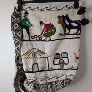 May include: Cream-colored textile bag with embroidered figures and scenes. The bag features a black strap with a geometric pattern and a decorative tassel. The embroidered scenes depict people, animals, and a hut, with a wavy border.