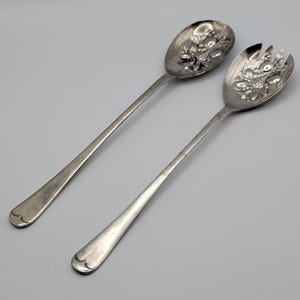 May include: A silver serving spoon and fork set. The spoon and fork have long, slender handles and decorative floral designs on the serving ends. The set is made of a shiny, reflective metal, likely silver or a silver-toned alloy.