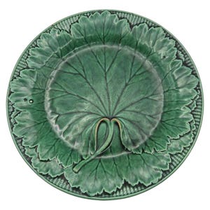 May include: A green ceramic plate with a leaf design. The plate features a large, central leaf motif with detailed veining, surrounded by a border of smaller leaves. The rim has a textured, woven pattern. The plate is a vibrant shade of green.
