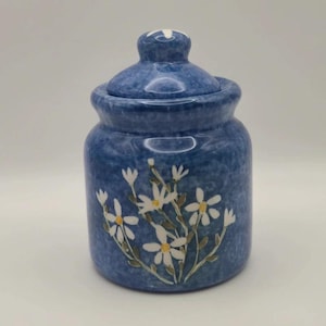 Könnte beinhalten: Ein blaues Keramikgefäß mit passendem Deckel, das eine gesprenkelte blaue Oberfläche und ein handgemaltes Design aus weißen Gänseblümchen mit gelben Mitten aufweist. Der zylindrische Behälter hat eine abgerundete Schulter und einen Deckel mit Knauf.
