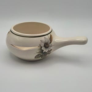 May include: A cream-colored ceramic soup bowl with a single handle, featuring a painted white flower and wheat stalk design on the side. The bowl has a distinct horizontal slit cut into the side wall and a thin dark green rim around the top edge.