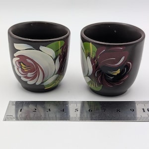 Peut inclure: Deux tasses en céramique brun foncé ornées de motifs floraux peints à la main en blanc, bordeaux et vert. Une règle en métal est placée devant les tasses, montrant que chaque tasse a un diamètre d'environ 5 cm au point le plus large.