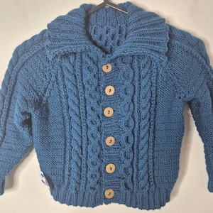 Peut inclure: Un cardigan tricoté bleu sarcelle profond avec un large col, des manches longues et un motif de torsades verticales. La fermeture avant se compose de six boutons en bois clair, ronds, disposés en une seule colonne au centre.