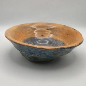 May include: A round ceramic bowl featuring a wide, flared rim with a warm tan and brown glaze. The interior base of the bowl transitions into a deep blue and grey tone, with visible circular throwing lines and subtle dark speckling throughout.