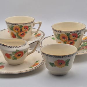 May include: A cream-coloured ceramic tea set featuring two teacups with matching saucers, a creamer pitcher on a saucer, and a small bowl. Each piece is decorated with painted orange and yellow flowers, green leaves, and a dark rim border.