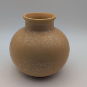 Könnte beinhalten: Eine kleine, runde, hellbraune Vase mit einem kurzen, schmalen Hals und einem breiten, kugelförmigen Körper. Die Oberfläche weist eine subtile, gesprenkelte Textur mit helleren, cremefarbenen Punkten auf dem matten, erdfarbenen Finish auf.