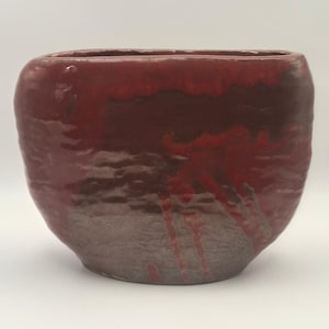 May include: A wide, oval-shaped ceramic vessel with a glossy finish. The upper portion features a vibrant, deep red glaze that drips down into a textured, dark brown base. The piece has a wide, open top and a rounded, organic silhouette.