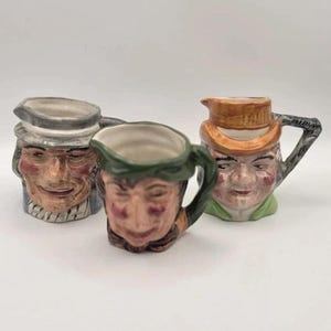 May include: Three ceramic character jugs featuring detailed facial expressions and painted features. The left jug wears a grey hat, the center jug has a green hat, and the right jug features an orange hat with a dark handle and green base.