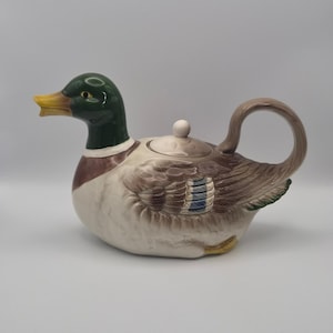 Op de afbeelding: Een keramische theepot in de vorm van een wilde eend met een donkergroene kop, een gele snavel en een lichaam met bruine, witte en blauwe veertjes. De theepot heeft een afneembaar deksel met een kleine knop en een gebogen handvat.