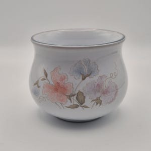 May include: A white ceramic bowl with a rounded shape and a smooth finish. The exterior features a delicate floral pattern in soft shades of pink, blue, and brown, depicting sweet pea flowers with green stems, leaves, and curling tendrils.