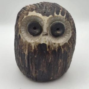 May include: A small, rounded ceramic owl figurine featuring a textured, dark brown and cream-coloured glaze. The owl has two large, dark, circular eyes set within a lighter, cream-coloured facial disc with fine, etched lines radiating outward.