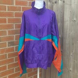 May include: A purple windbreaker jacket featuring a high collar, a full-length front zip, and long sleeves. The jacket is accented with horizontal and diagonal colour-blocked panels in bright teal and orange across the chest and sleeves.
