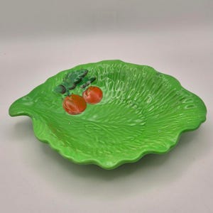 Puede incluir: Un cuenco de cerámica de color verde brillante con forma de hoja de lechuga grande y superficie texturizada. El cuenco presenta un diseño en relieve de dos pequeñas frutas redondas de color naranja rojizo con hojas verde oscuro.