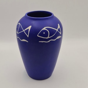 Könnte beinhalten: Eine matte kobaltblaue Vase mit bauchigem Körper und schmaler Öffnung. Die Außenseite ist mit handgemalten weißen Linien verziert, die zwei stilisierte Fische über wellenförmigen Linien darstellen und den oberen Teil des Gefäßes umschließen.