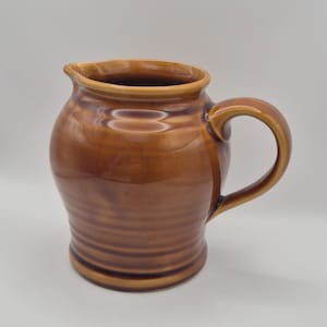 Peut inclure: Une cruche en céramique marron avec une anse et un bec verseur. La cruche a une finition brillante et une texture côtelée sur la moitié inférieure. La poignée est incurvée et fixée sur le côté. La cruche est de couleur marron chaud.