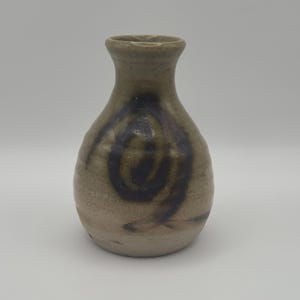May include: A small, bulbous ceramic vase with a narrow neck and a flared rim. The surface is a speckled light beige with a dark brown, hand-painted spiral design wrapping around the center of the body, featuring a smooth, glazed finish.