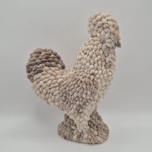 May include: A decorative rooster sculpture made from small seashells. The sculpture has a detailed design with a textured surface, in off-white and light brown hues. The rooster stands on a pedestal base.