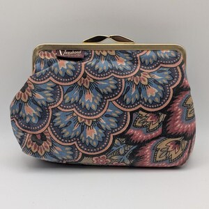 May include: A fabric coin purse featuring a repeating scalloped floral pattern in shades of blue, pink, and beige. The purse has a gold-toned metal frame with a kiss-lock closure and a small rectangular tag labeled Vagabond attached to the top.