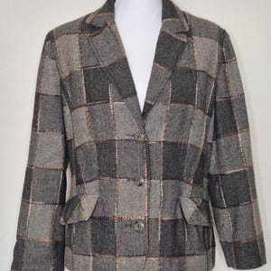 May include: A blazer featuring a dark grey and light grey herringbone pattern arranged in a large grid design. The garment includes a notched lapel, three front buttons for closure, and two flap pockets at the waist, all constructed from a textured fabric.