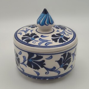 May include: A round ceramic lidded jar featuring a hand-painted floral pattern in dark blue and light blue on an off-white background. The lid includes a pointed, petal-shaped finial with matching blue decorative accents and circular lines.