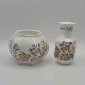 May include: Two white ceramic vases with floral and butterfly patterns. The larger vase is round with a hexagonal base. The smaller vase is a slender, elongated shape. Both vases have a glossy finish.