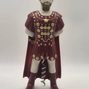 May include: A corkscrew designed as a standing figure wearing a dark red Roman-style tunic with gold-coloured circular accents and a matching cape. The figure has brown hair and a beard, with a silver-coloured metal spiral extending from the base.