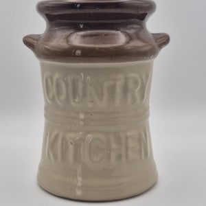 Könnte beinhalten: Ein zweifarbiger Keramiktopf mit dunkelbraun glasiertem Rand und beigefarbenem Körper. Das Gefäß verfügt über zwei kleine seitliche Griffe und den geprägten Schriftzug "COUNTRY KITCHEN" auf der Vorderseite. Der zylindrische Behälter hat eine weite Öffnung.