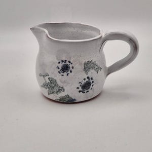 Può includere: Un piccolo brocca in ceramica bianca con manico e beccuccio. La brocca è decorata con motivi floreali blu scuro e verdi. La ceramica fatta a mano ha un aspetto rustico e strutturato, che suggerisce un design artigianale unico.