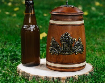 Könnte beinhalten: Eine braune Bierflasche und ein hölzerner Bierkrug mit einem dekorativen Metall-Emblem stehen auf einem Baumstumpf. Der Krug hat einen Deckel und ist mit einem Reliefdesign verziert. Die Gegenstände stehen vor einem Hintergrund aus grünem Gras.