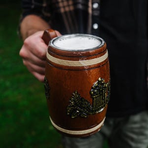 May include: A brown wooden beer mug with a decorative metal emblem and white foam. The mug has a barrel shape with white bands. The mug is being held by a person in a plaid shirt.