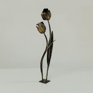 Puede incluir: Una escultura de metal bronce de dos tulipanes. Los capullos tienen detalles texturizados, y los tallos se curvan hacia arriba. La escultura está montada sobre una pequeña base cuadrada, y el diseño es elegante y minimalista.