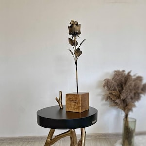 May include: A decorative side table with a black top and bronze-colored, branch-like legs. A metal rose sculpture is displayed on top, along with a small wooden block. A small bronze branch is also on the table.
