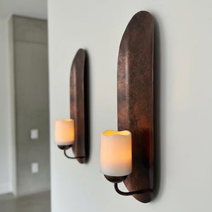 May include: Two wall-mounted candle holders with a copper finish. Each holder features a curved, vertical backplate and a black metal arm holding a white, battery-operated candle with a flickering, warm light. The design is minimalist and modern.