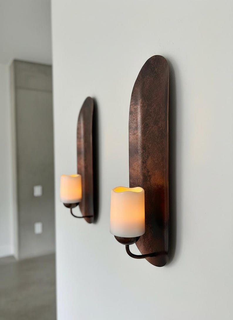 May include: Two wall-mounted candle holders with a copper finish. Each holder features a curved, vertical backplate and a black metal arm holding a white, battery-operated candle with a flickering, warm light. The design is minimalist and modern.