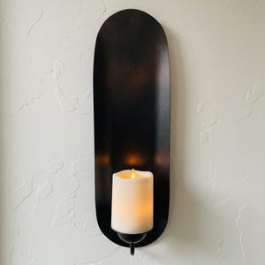 May include: A black metal wall sconce with a long, oval shape. It holds a white, battery-operated candle with a flickering yellow light. The sconce is mounted on a textured white wall, creating a warm, inviting ambiance.