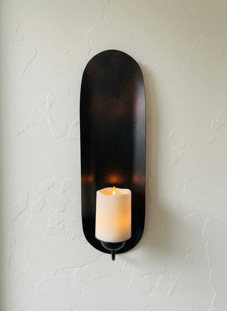 May include: A black metal wall sconce with a long, oval shape. It holds a white, battery-operated candle with a flickering yellow light. The sconce is mounted on a textured white wall, creating a warm, inviting ambiance.