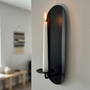 May include: A black metal wall sconce with a lit white candle. The sconce has a minimalist design with a curved backplate and a circular candle holder. The candle flame glows brightly against the dark background, creating a warm ambiance.