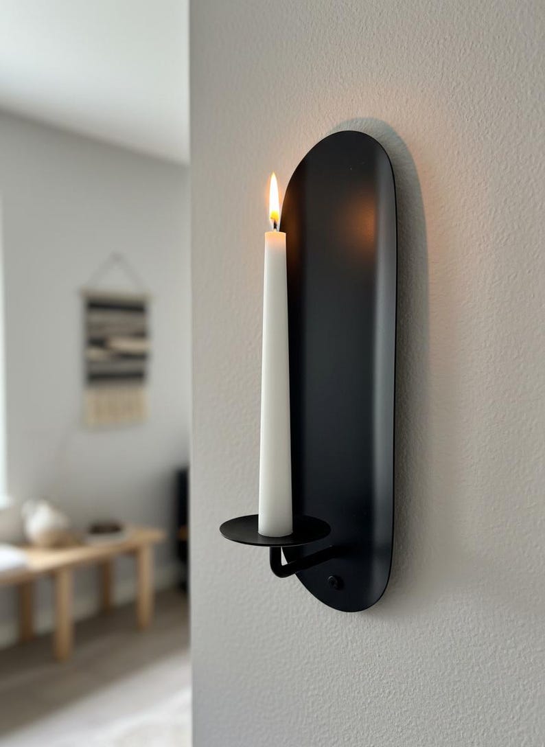 May include: A black metal wall sconce with a lit white candle. The sconce has a minimalist design with a curved backplate and a circular candle holder. The candle flame glows brightly against the dark background, creating a warm ambiance.