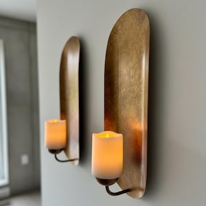 May include: Two wall sconces with a metallic gold finish. Each sconce features a curved, elongated backplate and a candle holder with a faux candle. The candles emit a warm, inviting glow, creating a cozy ambiance. The sconces are mounted on a light gray wall.