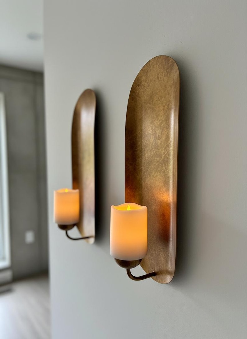 May include: Two wall sconces with a metallic gold finish. Each sconce features a curved, elongated backplate and a candle holder with a faux candle. The candles emit a warm, inviting glow, creating a cozy ambiance. The sconces are mounted on a light gray wall.