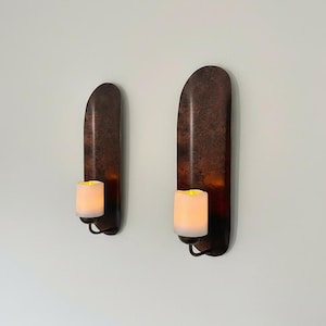 May include: Two wall-mounted candle holders with a dark, textured finish. Each holder features a curved, elongated design and holds a glowing, white candle. The holders are mounted on a white wall.