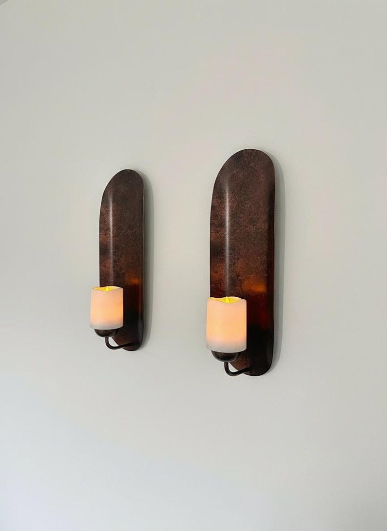May include: Two wall-mounted candle holders with a dark, textured finish. Each holder features a curved, elongated design and holds a glowing, white candle. The holders are mounted on a white wall.