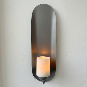 May include: A wall-mounted candle holder with a dark gray, elongated oval backplate. A white candle with a flickering yellow light sits in the center. The holder is designed for a modern aesthetic.