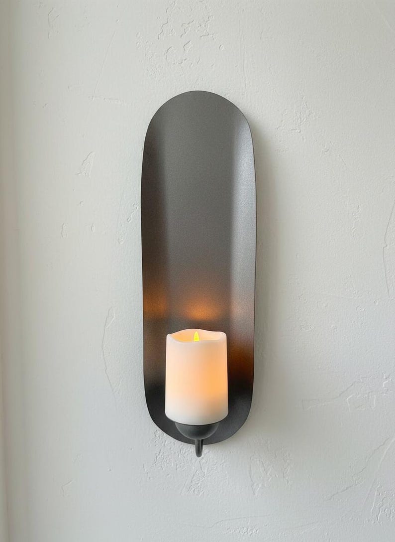 May include: A wall-mounted candle holder with a dark gray, elongated oval backplate. A white candle with a flickering yellow light sits in the center. The holder is designed for a modern aesthetic.