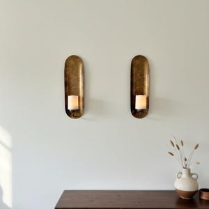 May include: Two antique brass wall sconces with a candle in each. The sconces have an oval shape and are mounted on a light-colored wall. A dark wooden table and a vase with dried plants are below.