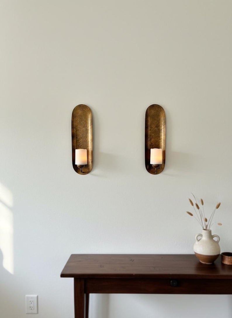 May include: Two antique brass wall sconces with a candle in each. The sconces have an oval shape and are mounted on a light-colored wall. A dark wooden table and a vase with dried plants are below.