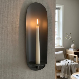 May include: A dark gray wall sconce with a lit, off-white candle. The sconce has a rounded, elongated shape and a small, circular base to hold the candle. The flame glows brightly against the dark background.